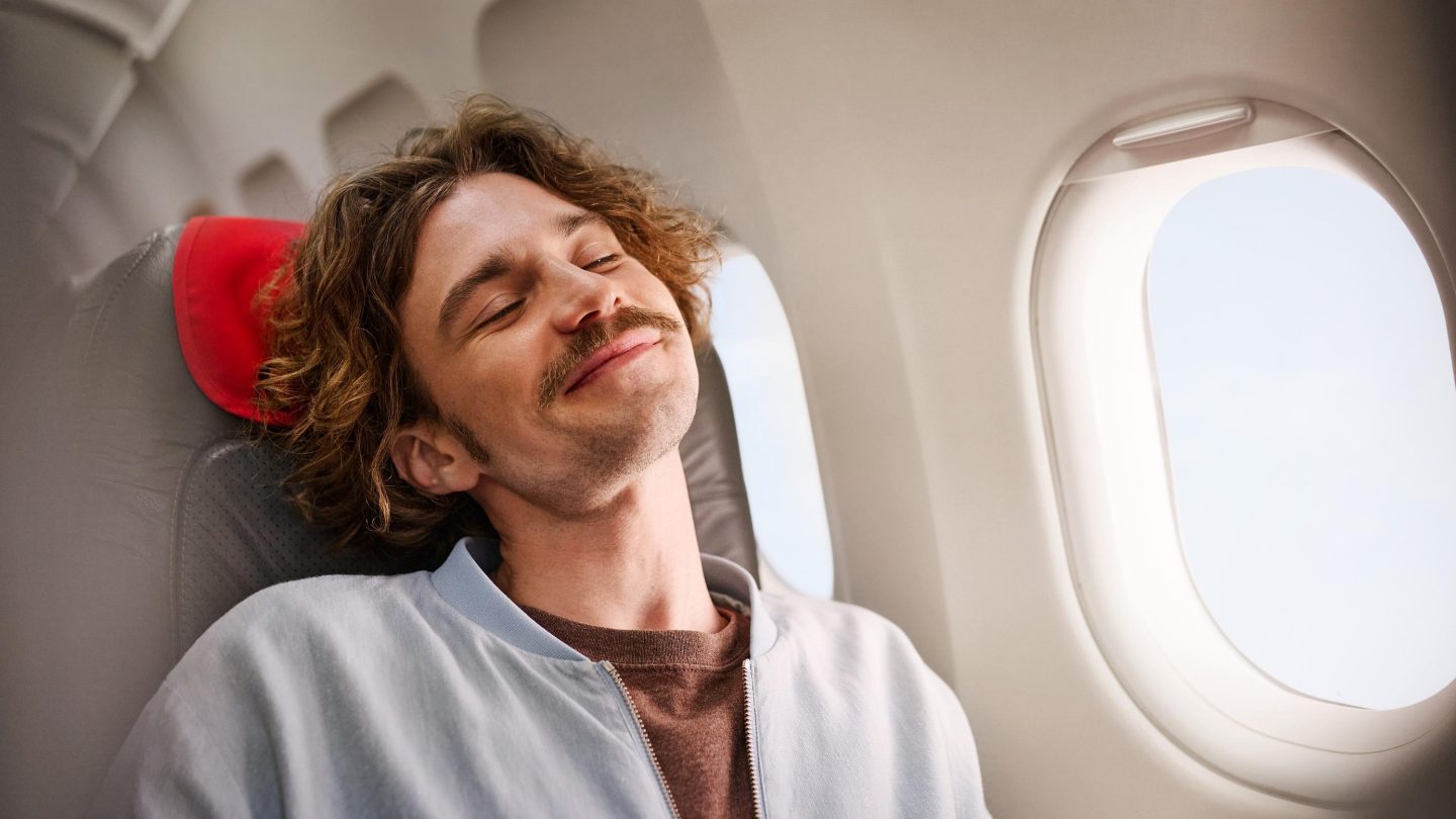 A smiling man relaxing comfortably in an airplane window seat with a red neck pillow.