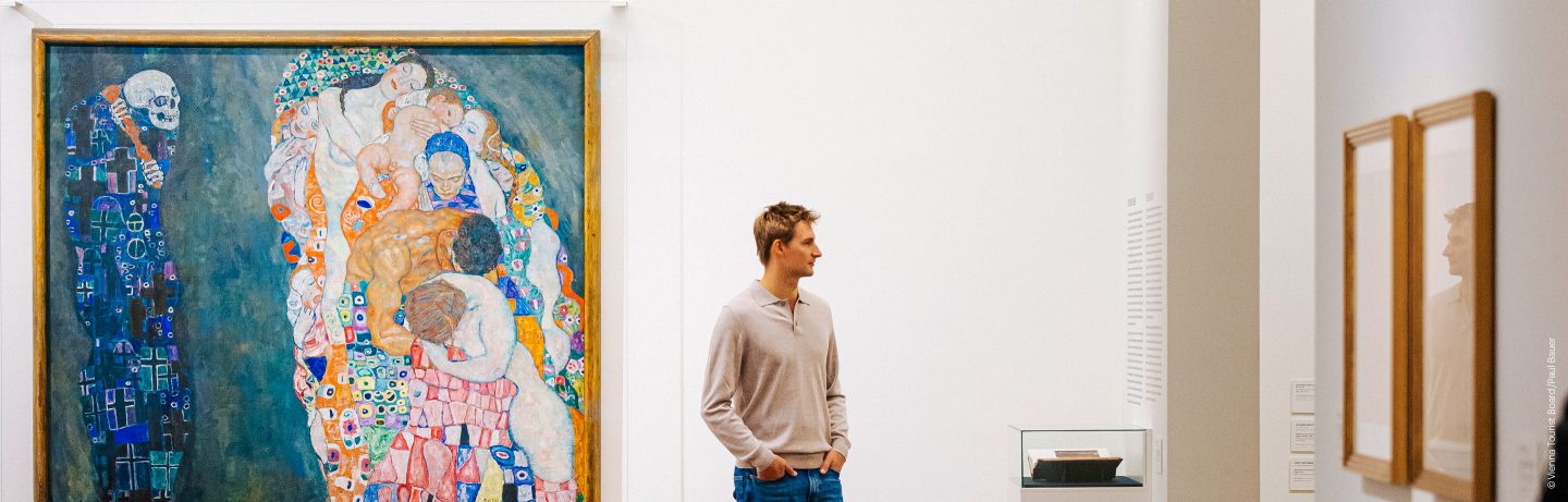 A man standing in a bright art gallery, looking at a large, colorful painting.