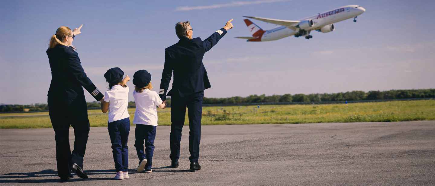 Two pilots and two children in uniform pointing at an Austrian Airlines plane flying overhead.